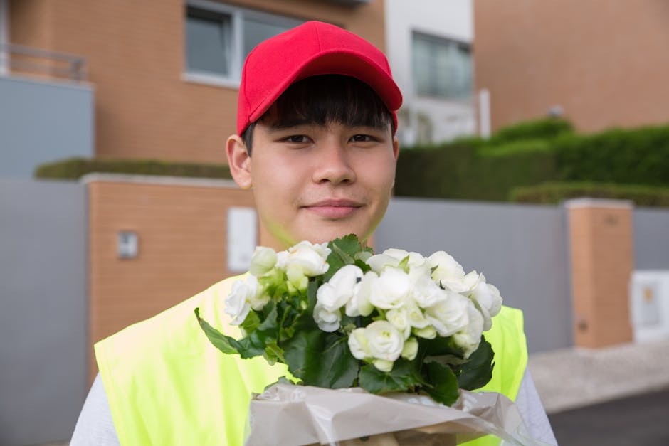 A young male flower delivery person wearing a red cap and high-visibility yellow vest is outdoors, standing in front of a modern residential building with brick and concrete exterior walls. He is hold