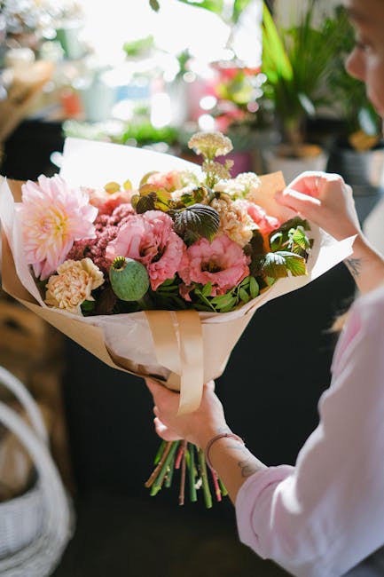 A person holding a beautifully arranged floral bouquet featuring large pink peonies, delicate cream carnations, and smaller white and purple flowers, complemented by lush green foliage. The bouquet is