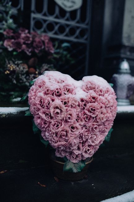 A heart-shaped floral arrangement composed of numerous pink roses with tightly packed, velvety petals, forming a dense, lush surface. The bouquet is displayed in a small container, resting on a dark s