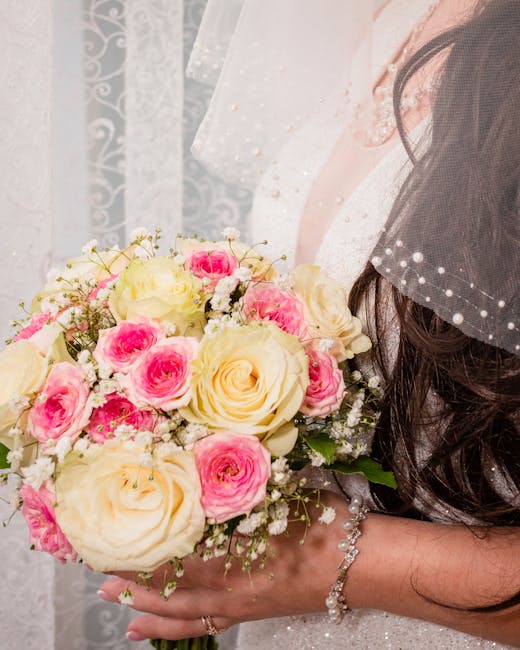 A close-up of a bridal bouquet featuring cream-colored roses and pink spray roses, accented with delicate baby's breath and green foliage. The bouquet is held by a bride dressed in a white wedding gow