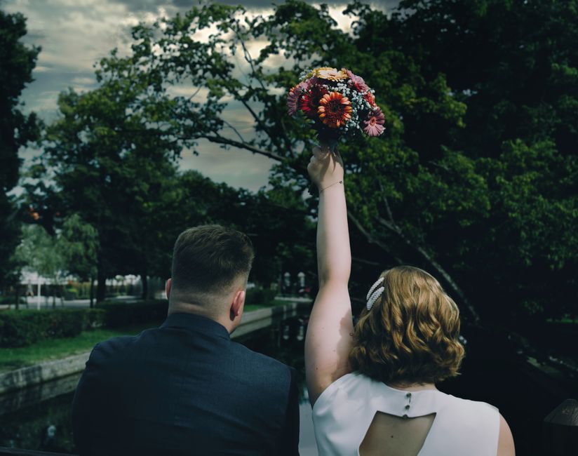 A woman holding up a bouquet of fresh flowers, featuring vibrant pink, orange, purple, and white blooms, arranged in a loose, natural style with visible stems and greenery, wrapped in clear and pastel