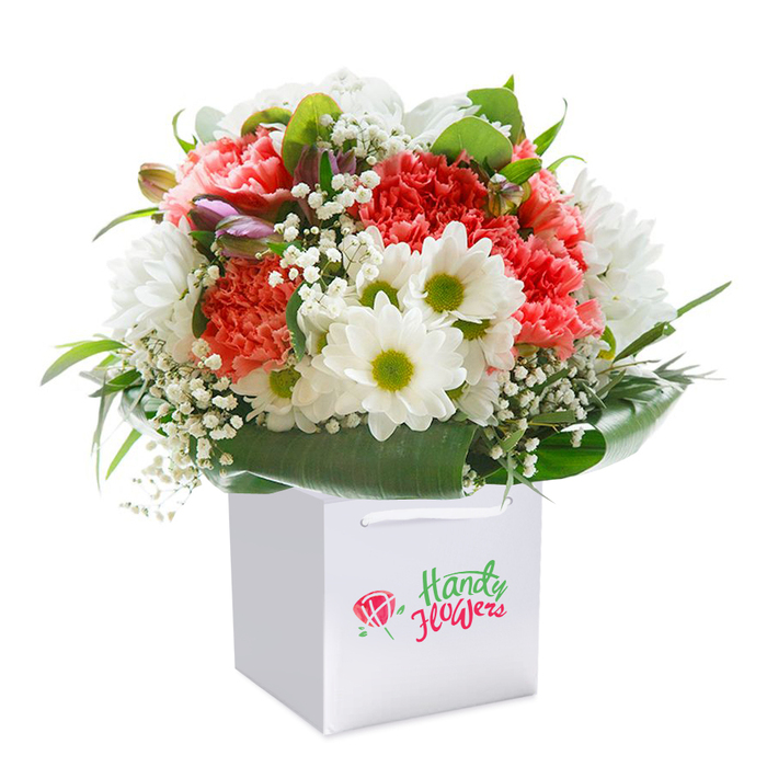 Against a flawless white backdrop, this festive floral arrangement stands out, carefully placed upright in a contemporary square gift bag-a hallmark of Upton Park's urban-chic floristry. The bouquet gathers an abundance of coral and rich red carnations, their frilled and layered petals imparting both texture and depth, joined by bright, white daisy-like flowers with luminous lime-green centers that radiate positivity. Flowing between the main blooms, baby's breath provides delicate, floating softness-a finishing touch akin to an early summer breeze in the parks of Upton Park. Notably, a few gently closed buds, tinged with purple and pink, adorn the arrangement's left flank, adding mystery, dimension, and the promise of new blossoms. The surrounding harmony of deep, broad, glossy green leaves creates an elegant natural wrap and a striking contrast to the lively floral palette. The arrangement's balanced, full, rounded silhouette reflects a practiced florist's hand and an understanding of both artistry and occasion. The sturdy white handles on the bag enhance the practical yet modern presentation, perfect for delivering a heartfelt surprise-be it birthday, anniversary, or a gesture for a neighbor-anywhere from Green Street Market to the quieter corners of Upton Park. Every detail, from texture to color, reflects the vibrant diversity and community connection that defines this East London neighborhood.